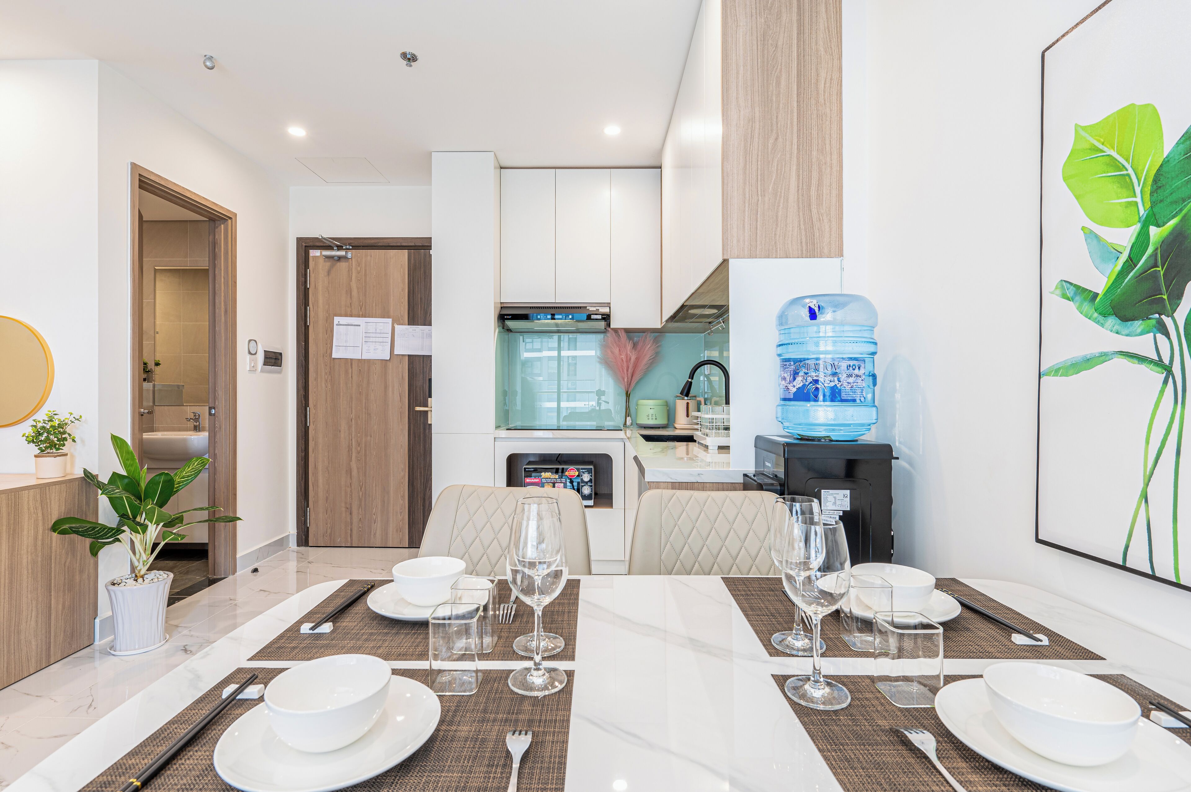 Un comedor moderno con una mesa preparada para cuatro, una cocina al fondo y un dispensador de agua en la pared.