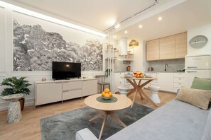 A modern living room with a flat-screen TV, a dining table with a bowl of fruit, a sofa, and a kitchen area with cabinets and a clock. - Bright Studio with Balcony | Ideal for Remote Work (Łódź)