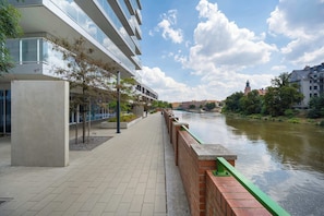 Une promenade au bord de la rivière, avec un bâtiment moderne, un pont et une tour qui se dresse au loin.