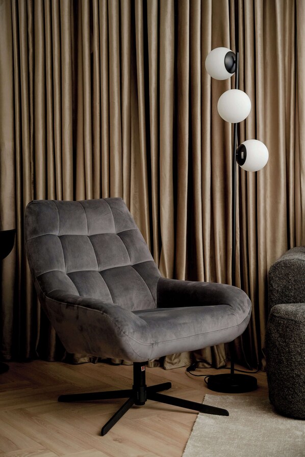 A modern chair with a tufted backrest and a swivel base, positioned in front of a curtain backdrop and a floor lamp with three spherical lights.