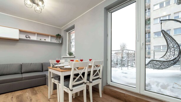 A modern living room with a sofa, dining table, chairs, and a large window with a view of a snowy outdoor area. - Cozy Studio with Balcony | Parking in Garage (Łódź)