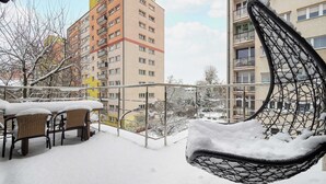 A balcony with a hanging chair covered in snow, a table with chairs, and a view of multi-story buildings. - Cozy Studio with Balcony | Parking in Garage (Łódź)