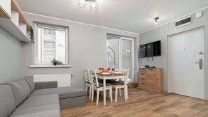A modern living room with a grey sofa, a dining table with chairs, a TV mounted on the wall, and a wooden cabinet. - Cozy Studio with Balcony | Parking in Garage (Łódź)
