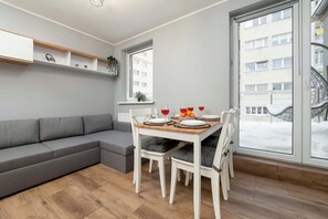 Ein modernes Wohnzimmer mit einem grauen Sofa, einem Esstisch mit Stühlen, einem Fenster mit Blick auf ein Gebäude und einem Regal mit Dekorationen.