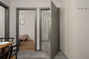 A hotel room with a bed, a small table, a chair, a shower, and a door leading to another room.