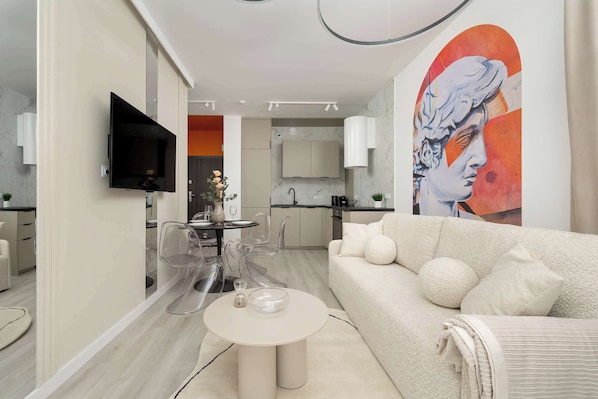 A modern living room with a sofa, coffee table, and clear chairs. A TV is mounted on the wall, and a kitchen is visible in the background.