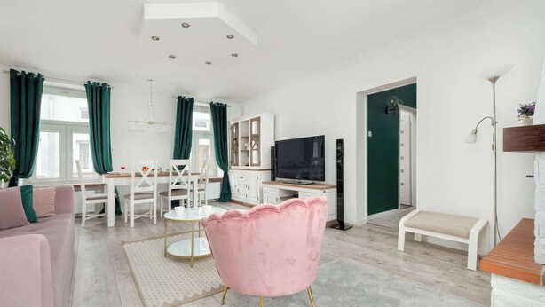 A modern living room with a pink armchair, a dining table with chairs, a TV, and a staircase leading to another level. - Spacious Apartment in the City Center | Balcony | 2 Bedrooms (Łódź)