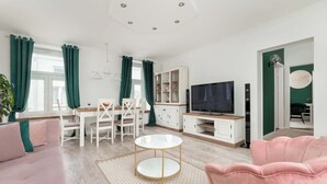A modern living room with a sofa, a pink armchair, a dining table with chairs, a TV, and a cabinet. - Spacious Apartment in the City Center | Balcony | 2 Bedrooms (Łódź)