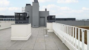 Eine Dachterrasse mit weißer Brüstung, einem kleinen runden Tisch und einem grauen Gebäudebau.