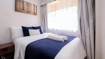 Ein ordentlich bezogenes Bett mit weißer und blauer Bettwäsche, ein Nachttisch aus Holz, ein Bild an der Wand und ein Fenster mit Vorhängen.