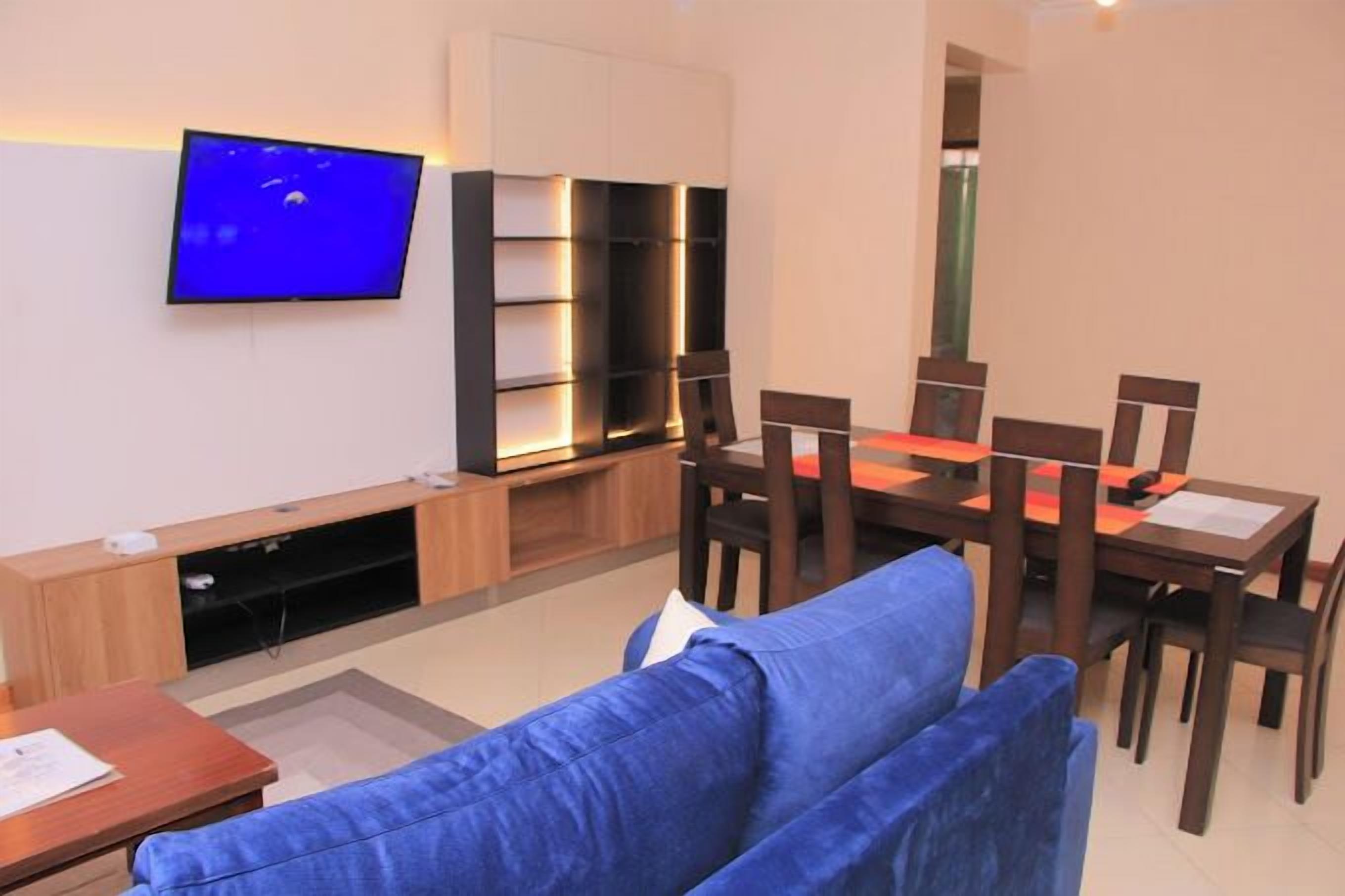 Una sala de estar moderna con un televisor de pantalla plana montado en la pared, un mueble de entretenimiento de madera, un sofá azul y una mesa de comedor con sillas.