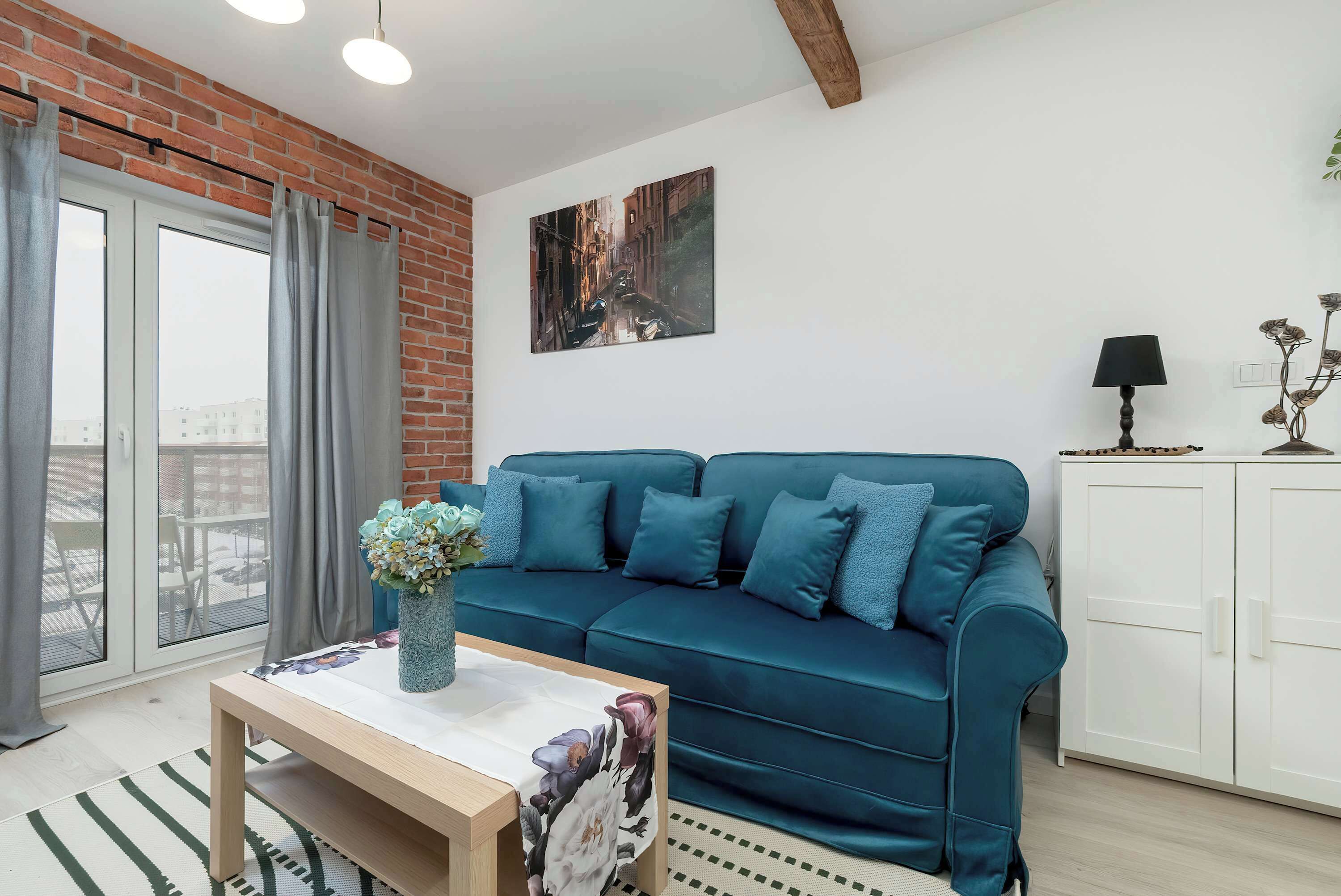 Una sala de estar con un sofá azul, una mesa de centro de madera, un florero con flores, un cuadro en la pared y un mueble blanco.