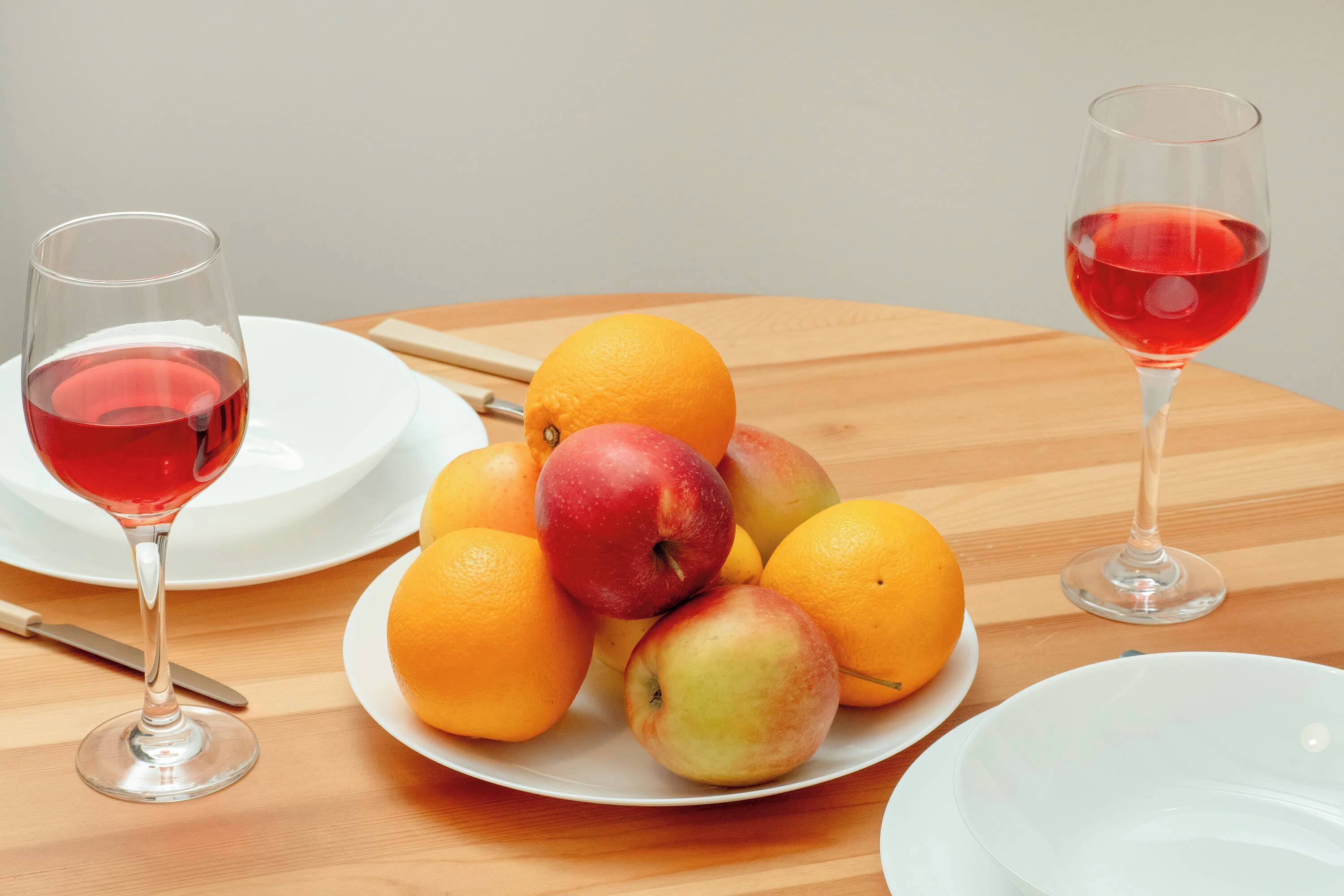 A wooden table with two white plates, one with oranges and apples, and two wine glasses with red wine.