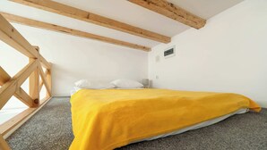 Een slaapkamer met een bed belegd met een gele deken, houten balken in het plafond en een raam aan de linkerkant.