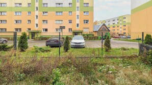 Een meerlaags woongebouw met een geelgroene gevel, een omheind parkeerterrein en een klein gebouw met een groen dak.