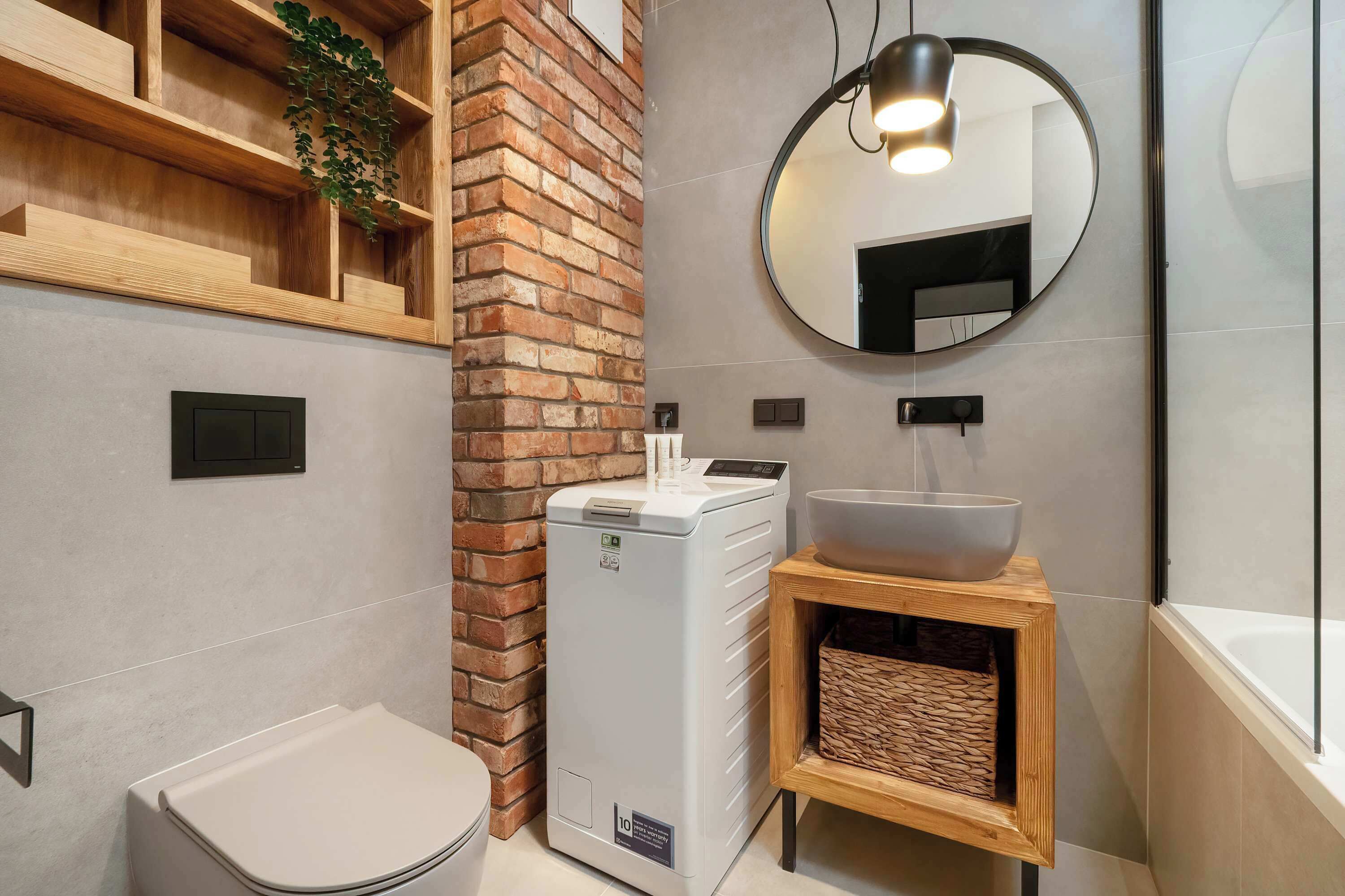 Un baño moderno con pared de ladrillo decorativa, calefacción integrada, espejo redondo y tocador de madera con lavabo tipo cuenco.