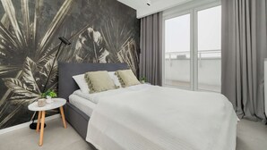 Ein modernes Schlafzimmer mit einem großen Bett, einem kleinen runden Tisch, einer Pflanze und Blick ins Freie durch ein großes Fenster.