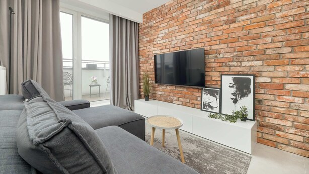 Ein modernes Wohnzimmer mit Backsteinwand, Flachbildfernseher, grauen Sofas und einem kleinen runden Tisch.