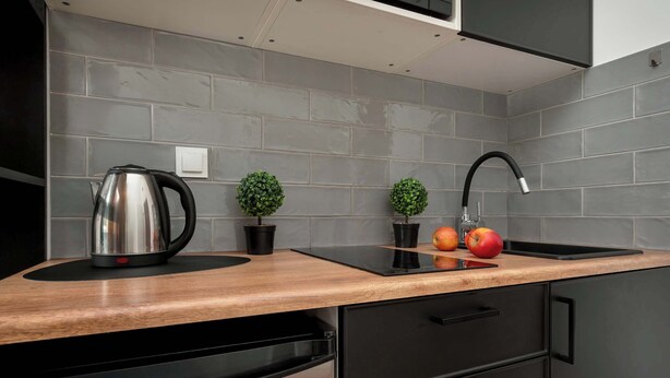 Una cocina moderna con baldosas grises, encimera de madera, un hervidor, dos plantas en macetas y un frutero.