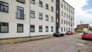 Un edificio de varios pisos con fachada blanca y marcos de ventanas negros. Varios autos están estacionados al frente, incluyendo un auto rojo y uno negro.