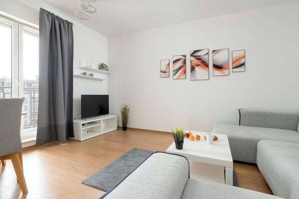 A modern living room with a grey sofa, a white coffee table, and a flat-screen TV on a white stand. There are abstract wall art pieces and a potted plant on the coffee table.