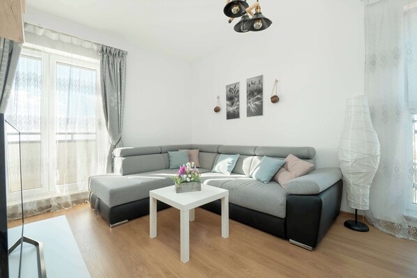 Ein modernes Wohnzimmer mit einem grauen Ecksofa, einem weißen Couchtisch und einem großen Fenster mit durchsichtigen Vorhängen.