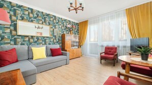 A modern living room with a grey sofa, red and yellow cushions, a wooden cabinet, a red armchair, a round wooden table, and a large window with yellow curtains.