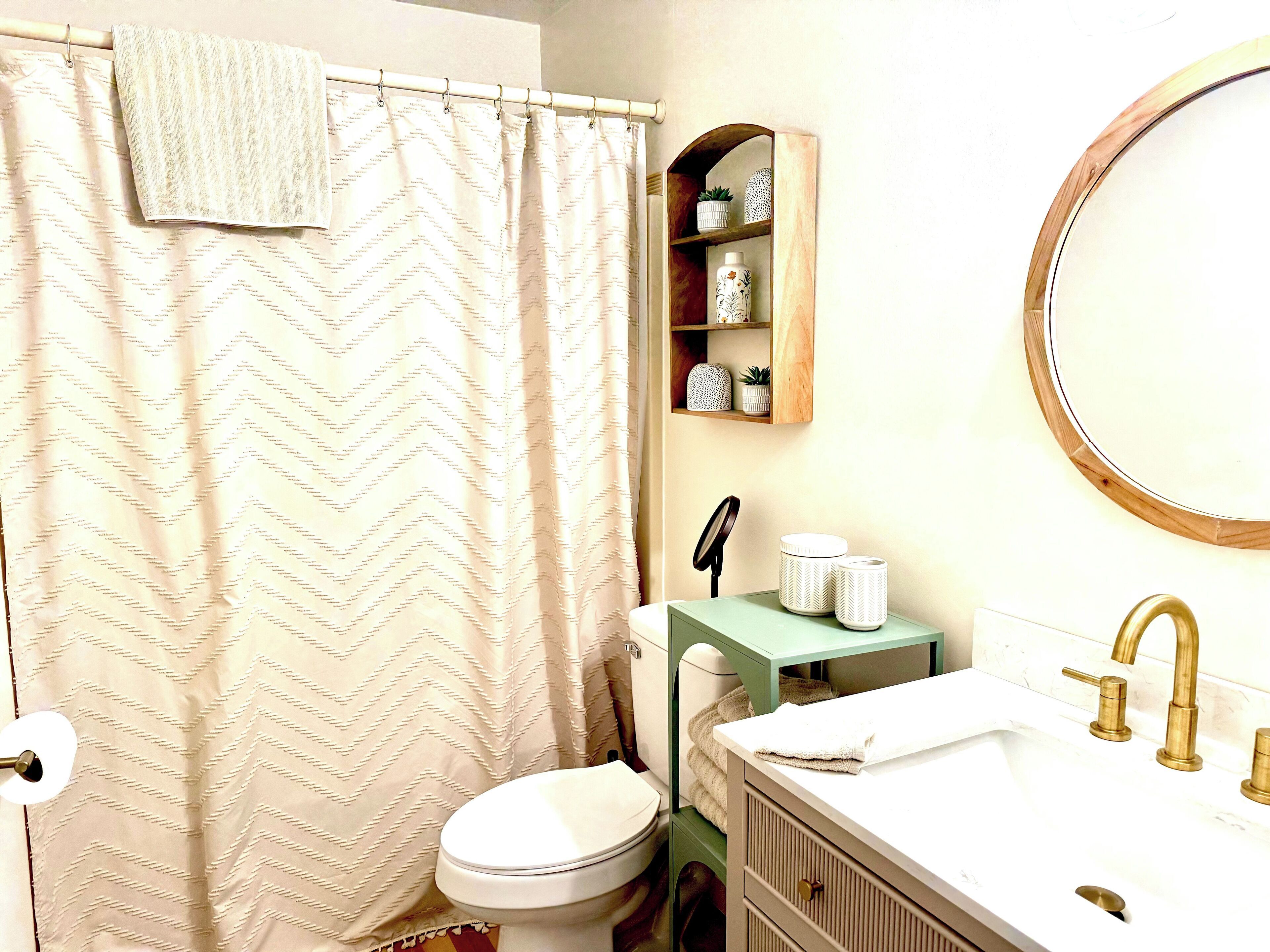 Un baño con inodoro, lavamanos con espejo, cortina de ducha y estante con objetos decorativos.
