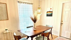 A dining area with a wooden table, four chairs, a vase with dried plants, a hanging light fixture, and a painting on the wall. - Southern Charm Under the Oaks - Pet Friendly (Ocean Springs)
