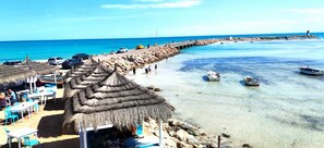 Una playa con construcciones de techo de paja, aguas poco profundas y cristalinas, y un muelle que se adentra en el mar.