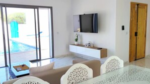 En moderne stue med et glasbord, hvide stole, en sofa, et tv ophængt på væggen og en skydedør af glas, der fører til et udendørs område med en pool.