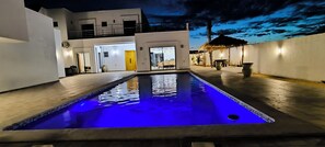 A swimming pool with a blue-lit interior, surrounded by a modern building with a balcony and a thatched-roof structure.