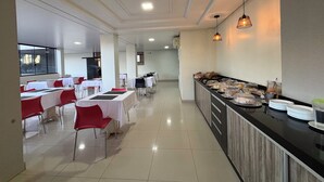 Un'ampia sala da pranzo con numerosi tavoli e sedie, un'area buffet con diverse pietanze e una cucina moderna sullo sfondo.
