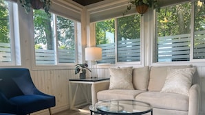 A room with a blue armchair, a beige sofa with patterned pillows, a glass coffee table, and a small side table with a lamp and plant. - Ravenwood (Niagara-on-the-Lake)