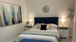 A bedroom with a large bed, two bedside tables, a bench, a painting, and a decorative mirror. - Ravenwood (Niagara-on-the-Lake)