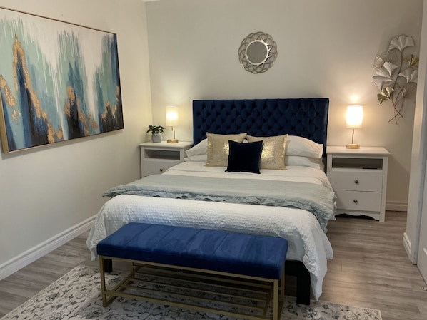 A bedroom with a large bed, two bedside tables, a bench, a painting, and a decorative mirror. - Ravenwood (Niagara-on-the-Lake)