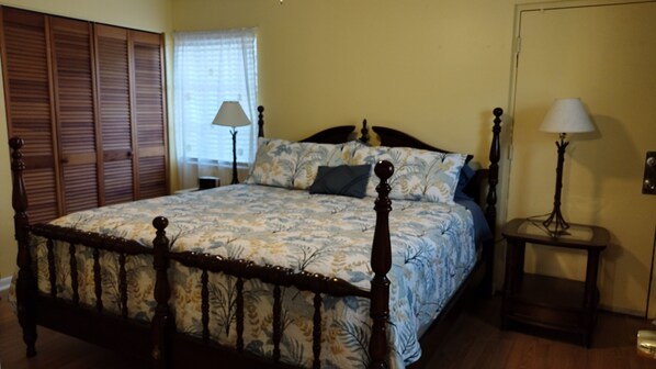 A bedroom with a wooden four-poster bed, a floral bedspread, a nightstand with a lamp, and a window with blinds. - Three Sisters Cottages (Crystal River)
