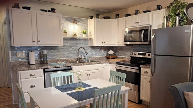 A compact kitchen with white cabinets, a microwave, a refrigerator, and a dining area with a table and chairs. - Three Sisters Cottages (Crystal River)