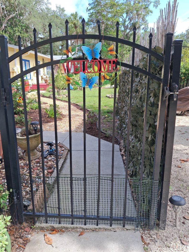 A gated entrance with a 'WELCOME' sign, a butterfly decoration, and a pathway leading to a house.