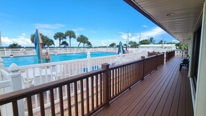 Una terraza de madera con piscina, barandas blancas y palmeras al fondo.
