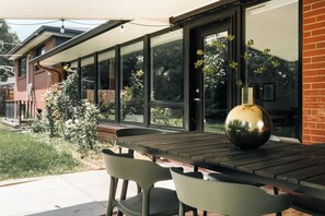 Een terras buiten met een tafel, stoelen, een groot raam en een vaas met bloemen.