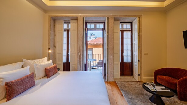 Habitación de hotel con una cama grande, una silla roja, una mesita y una puerta abierta que da a un espacio al aire libre.