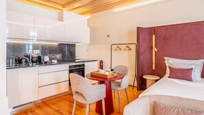 Quarto de hotel moderno com cozinha, área de jantar com duas cadeiras, cama com cabeceira vermelha e uma pequena mesa de cabeceira.