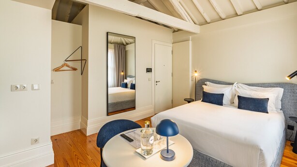 A hotel room with a bed, a round table with a bottle and remote, a mirror, and a coat hanger.
