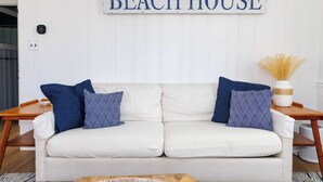 Un sofá blanco con almohadones de diseño azul y blanco, una mesita de madera y un letrero en la pared que dice 'BEACH HOUSE'.