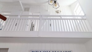 Un interior blanco con un cartel que dice 'BEACH HOUSE', una escalera y una lámpara con diseño geométrico.