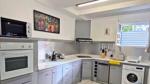 Een moderne keuken met een ingebouwde oven, magnetron en vaatwasser. Er is een raam met jaloezieën, een spoelbak en een prullenbak.