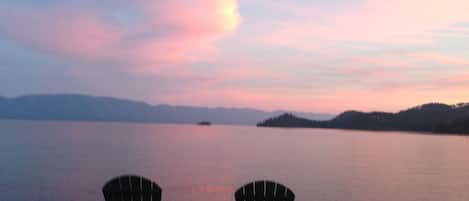 Two Adirondack chairs facing a lake with a sunset.