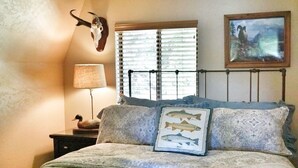 Een slaapkamer met een bed, een lamp, een aan de muur bevestigd schedel, een ingelijst kunstwerk en een raam met zonwering.
