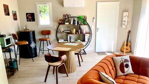 Ein modernes Wohnzimmer mit einem runden Holztisch, zwei braunen Sesseln, einem orangefarbenen Sofa mit Kissen, einem Bücherregal und einer Gitarre.